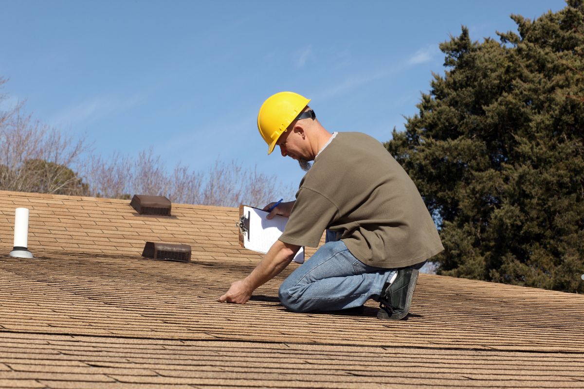 A worker in a yellow hard hat kneels on a textured roof, inspecting it while holding a clipboard and a pen under a clear blue sky.