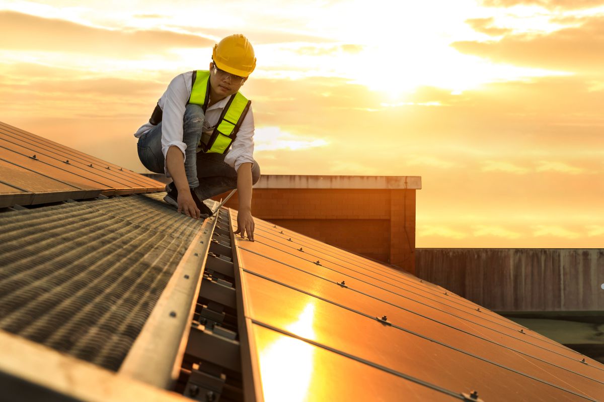A worker in a yellow safety vest examines solar panels at sunset, with the sky painted in warm hues of orange and gold.