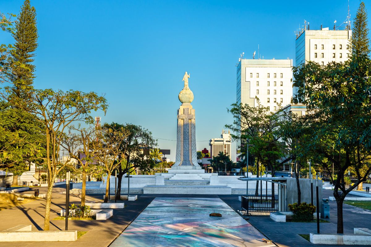 A sunny urban plaza featuring a tall monument topped with a cross, surrounded by trees and modern buildings.